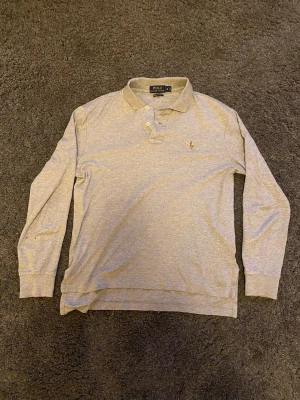 Grå långärmad piké från Ralph Lauren - Säljer en stilren grå långärmad pikétröja från Polo Ralph Lauren i mjuk Pima-bomull. Klassisk krage, knäppning med tre knappar och den ikoniska broderade loggan på bröstet. Perfekt till jeans eller chinos.