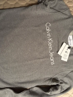 Grå sweatshirt från Calvin Klein Jeans - Snygg grå sweatshirt från Calvin Klein Jeans med logga tryckt i vitt på bröstet. Tröjan har rund halsringning och ribbade muddar vid ärmslut och nederkant. Perfekt för en avslappnad och trendig look.