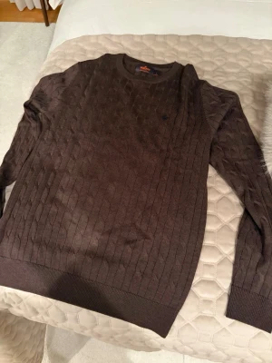 Brun kabelstickad tröja Morris - Snygg brun kabelstickad tröja från Morris med rund halsringning och diskret logga broderad på bröstet. Tillverkad i mjukt material som passar perfekt till höst och vinter. Klassisk modell med ribbade muddar vid ärmslut och nederkant.