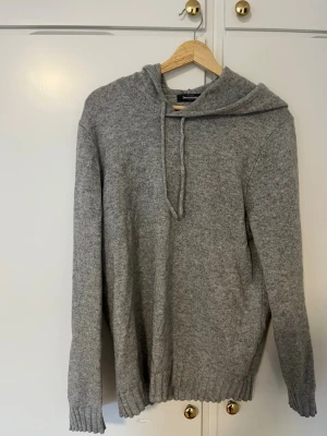 Grå stickad hoodie med huva - Mysig grå stickad hoodie med huva och dragsko. Tröjan har långa ärmar, ribbstickade muddar och bred ribbkant nedtill. Perfekt för lager-på-lager och chill dagar. Materialet ser ut att vara mjukt och värmande ullblandning.L passar M!