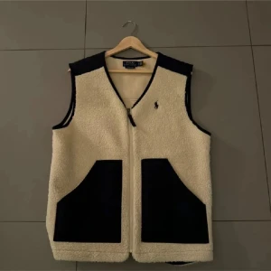 Beige fleeceväst från Polo Ralph Lauren - Snygg beige fleeceväst från Polo Ralph Lauren med svarta detaljer och två stora fickor framtill. Västen har dragkedja och klassisk logga broderad på bröstet. Perfekt lager-på-lager-plagg för kyligare dagar. Är som helst ny, använd 3-4 gånger.