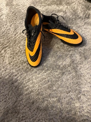 Nike orange-svarta fotbollsskor - Säljer ett par snygga fotbollsskor från Nike i orange och svart med klassisk swoosh-logga. Skorna har snörning och ovandel i syntetmaterial med textur för bättre bollkontroll. Perfekta för dig som vill sticka ut på planen!