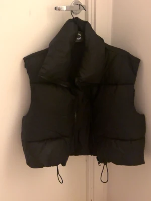 Svart cropped dunväst med hög krage - Snygg svart dunväst med cropped passform och hög krage. Västen har dragkedja framtill och justerbara snören i nederkant för en cool look. Perfekt att slänga över en hoodie eller tröja för extra värme. Enkel och stilren design som passar till det mesta.