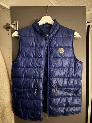Blå dunväst från Moncler - Snygg blå dunväst från Moncler med ståkrage och två dragkedjeförsedda fickor framtill. Västen har quiltad design och ett broderat Moncler-märke på bröstet. Perfekt för lager på lager och ger en sportig vibe.