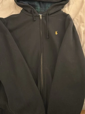Svart zip hoodie från Polo Ralph Lauren - Svart hoodie med dragkedja från Polo Ralph Lauren. Klassisk design med huva och justerbara snören, samt det ikoniska gula broderade logotypen på bröstet. Tillverkad i mjukt bomullsmaterial som är skönt att ha på sig. Perfekt för en avslappnad och stilren look.