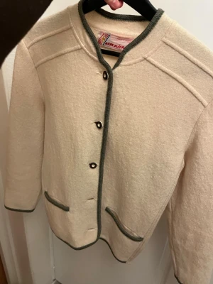 Beige kofta från Spieth & Wensky - Stilren beige kofta från Spieth & Wensky med gröna kanter och dekorativa knappar framtill. Koftan har rund halsringning, två fickor och är tillverkad i ett mjukt, ulligt material. Perfekt för dig som gillar klassisk och tidlös stil.