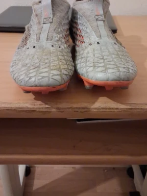 Grå-orange Puma fotbollsskor - Säljer ett par gråa fotbollsskor från Puma med orange detaljer och sula. Skorna har ett mönstrat yttre i syntetmaterial och meshpaneler på sidorna för extra ventilation. Designen är modern och strömlinjeformad, perfekt för dig som vill sticka ut på planen.