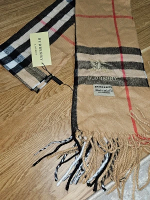 Beige rutig halsduk från Burberry - Klassisk beige halsduk från Burberry med det ikoniska rutmönstret i svart, vitt och rött. Halsduken har fransar i ändarna och en broderad Burberry-logga. Tillverkad i mjukt material som känns lyxigt och stilrent. Perfekt accessoar för att lyfta din outfit. helt ny