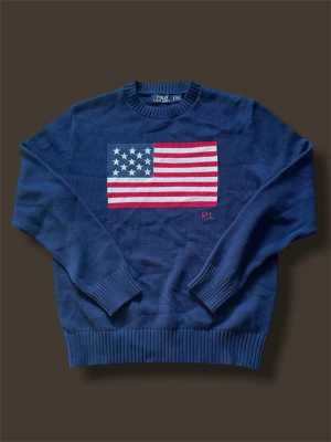 Mörkblå stickad tröja Polo Ralph Lauren - Mörkblå stickad tröja från Polo Ralph Lauren med stor amerikansk flagga på bröstet. Tröjan har rund halsringning och ribbade muddar vid ärmslut och nederkant. Klassisk och tidlös design med RL-broderi under flaggan. Det står storlek M men den är ganska liten i storlek så passar bättre som storlek S. Modellen är 180 lång och väger 65.