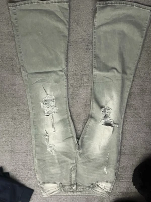 Grå slitna bootcut jeans - Säljer ett par grå bootcut jeans med slitningar och hål på framsidan av benen. Jeansen har klassisk femficksdesign och vida ben. Perfekta för dig som gillar en avslappnad och trendig stil och väldigt stretchiga.