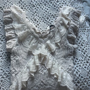 Beige spetsblus med volang från Zara - Supersöt beige blus från Zara med v-ringning och massor av spetsdetaljer. Blusen är ärmlös och har volanger längs axlar och framtill, vilket ger en romantisk och trendig look. Perfekt för dig som gillar unika och detaljerade toppar!