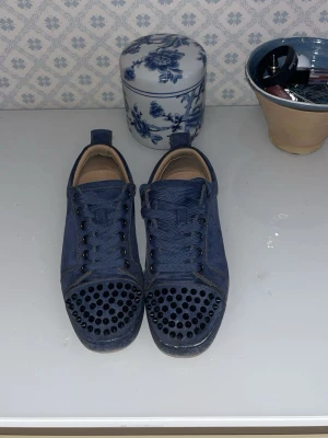 Blå Louboutin skor med nitar på tån - Snygga blå sneakers i mocka med coola svarta nitar på tån och matchande blå skosnören. Skorna har lågt skaft och svarta metalldetaljer vid snörningen. Perfekta för dig som vill sticka ut med din stil.