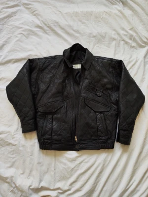 Lammskinnjacka - Snygg svart lammskinnjacka med quiltat mönster, dragkedja framtill och flera fickor med lock. Jackan har klassisk krage och ribbade muddar i midjan. Perfekt för dig som vill ha en tidlös och cool look.