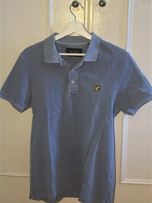 Blå pikétröja från Lyle & Scott - Klassisk blå pikétröja från Lyle & Scott med korta ärmar och krage. Tröjan har knappslå framtill och den ikoniska gula loggan broderad på bröstet. Perfekt för en avslappnad och stilren look.