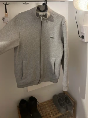 Grå zip-hoodie från Lacoste - Snygg grå zip-hoodie från Lacoste med klassisk krokodillogga på bröstet. Tröjan har dragkedja hela vägen, två fickor fram och ribbade muddar. Tillverkad i mjukt bomullsmaterial som är skönt mot huden. Perfekt för en avslappnad och stilren look.