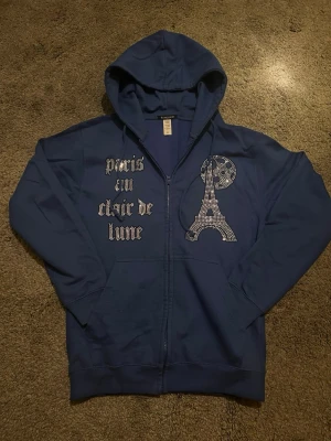 Rhinestone hoodie - Blå zip-hoodie från BLINGLUCID med huva och fickor. Framsidan har texten 'paris en clair de lune' i silvrigt tryck och en glittrig Eiffeltorn-design i strass. Tillverkad i bomull och polyester, perfekt för en trendig och avslappnad look.