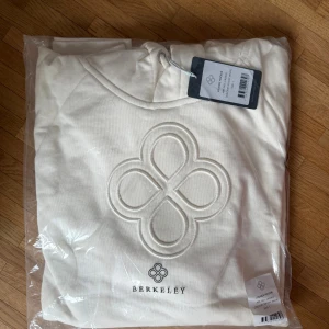 Offwhite hoodie från Berkeley - Snygg offwhite hoodie från Berkeley med stor broderad logga på bröstet och matchande tryck under. Hoodien har huva med dragsko och är tillverkad i mjukt material som passar perfekt för en chill och avslappnad stil.