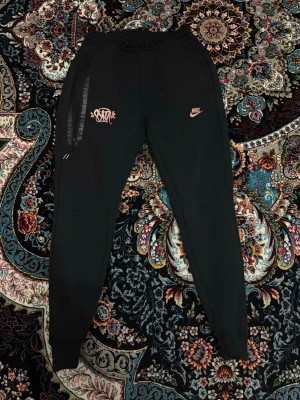 Syna World Nike Byxor - Säljer ett par svarta mjukisbyxor från Nike med rosa logga och tryck på vänster ben. Byxorna har ribbade muddar vid bensluten, dragsko i midjan och en ficka med dragkedja på sidan. Sköna och stilrena, perfekta för chill eller träning.