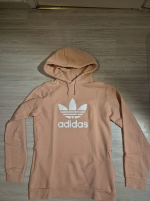 Beige hoodie från Adidas Originals - Snygg beige hoodie från Adidas Originals med stor vit logga framtill. Klassisk modell med huva och dragsko, samt ribbade muddar vid ärmslut och nederkant. Tillverkad i mjukt bomullsmaterial som är skönt mot huden. Storleken är xs men skulle säga att den också kan passa s. Färgen är typ beige/rosa 