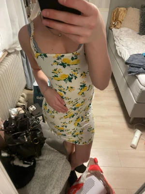 Vit blommig kortklänning - Söt vit kortklänning med gult och grönt blommönster. Klänningen har smala axelband och en figurnära passform. Perfekt för varma dagar och somriga tillfällen. Kommer från boohoo i storlek XS. 