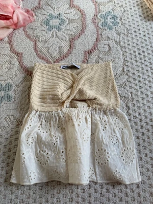 Beige och vit broderad topp från Zara - Supersöt topp från Zara i beige och vitt. Överdelen är virkad med twistad detalj och cut-out framtill, medan nederdelen är i broderad bomull med blommönster. Axelbandslös och perfekt till sommaren för en chill och trendig look. Populär topp 
