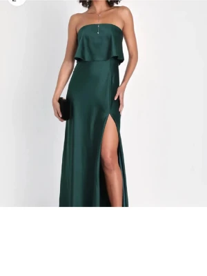 Mörkgrön axelbandslös balklänning - Snygg mörkgrön axelbandslös långklänning i satinliknande material med volang upptill och hög slits på ena sidan. Perfekt för bal eller festliga tillfällen. 