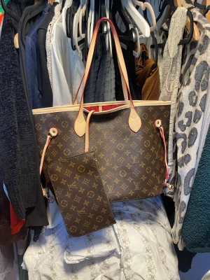 Louis Vuitton Neverfull Monogram väska - Säljer en klassisk Louis Vuitton Neverfull handväska i brunt monogramcanvas med beige läderdetaljer och rödrandigt innerfoder. Väskan har långa axelband, guldiga metalldetaljer och medföljande clutch. Perfekt för dig som gillar lyxig och tidlös design.