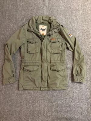 Grön vindjacka från Superdry - Snygg militärgrön vindjacka från Superdry med fyra stora fickor framtill, patch på ärmen och dragkedja med tryckknappar. Jackan är i helt nytt skick finns inga defekter, jackan är även sällsynt. Kom dm vid frågor✅