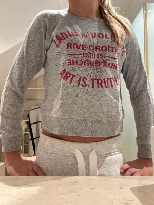 Grå sweatshirt Zadig & Voltaire - Snygg grå sweatshirt från Zadig & Voltaire med röd text och tryck framtill. Tröjan har rund halsringning, ribbade muddar och avslappnad passform. Perfekt till jeans eller mjukisbyxor för en chill look. Några paljetter som lossnat