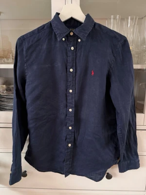 Mörkblå skjorta från Polo Ralph Lauren - Snygg mörkblå skjorta från Polo Ralph Lauren med klassisk krage och knappar framtill. Skjortan har lång ärm och är tillverkad i mjuk bomull. På bröstet finns den ikoniska röda Polo-loggan broderad. 