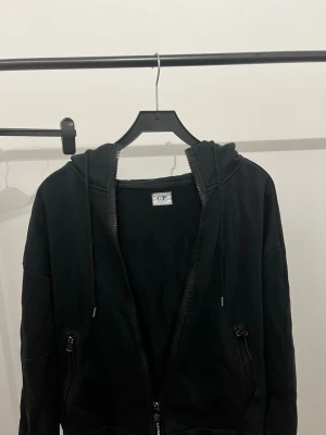 Svart hoodie med dragkedja från CP company - Svart hoodie från CP med dragkedja framtill, justerbar huva med snören och två fickor med dragkedja. Tillverkad i mjukt bomullsmaterial och har ribbade muddar vid ärmslut och nederkant. Perfekt för en avslappnad och stilren look.