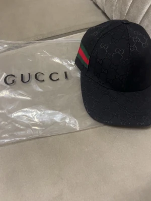 Svart Gucci keps med GG-mönster - Snygg svart keps från Gucci med det klassiska GG-monogrammet över hela kepsen. På sidan finns en grön och röd rand som ger en ikonisk touch. Kepsen är tillverkad i tyg och har en böjd skärm. Perfekt för dig som vill ha en exklusiv och trendig accessoar.
