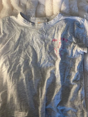 Ljusgrå t-shirt från Gina Tricot - Ljusgrå t-shirt från Gina Tricot med korta ärmar och rund hals. På bröstet finns en rosa broderad stjärna och texten 'MA CHÉRIE'. Perfekt för en avslappnad och trendig look.