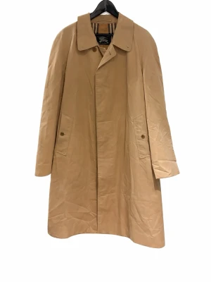 Vintage Burberry Trench Coat/ Rock - Klassisk vintage Trench Coat / Rock från Burberry. Tillverkad i England.                                  Herrmodell. Tillverkad under en tid då Burberry inte inkluderade storlekslappar i kläderna.              Uppskattad storlek: L                                                 Mått: Bröst: ca 116cm. Rygglängd: ca 103cm.         Plagget är i bra skick med normalt bruksslitage. Finns en liten fläck under höger ficka på framsidan (se bilder)