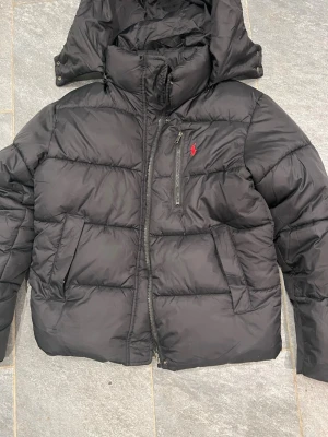 Svart dunjacka från Ralph Lauren - Aldrig använd!!! Säljer en svart dunjacka från Ralph Lauren med huva och dragkedja. Jackan har en bröstficka med dragkedja och den klassiska röda loggan på bröstet. Feta kanaler och två sidofickor gör den både snygg och praktisk. Perfekt för kalla vinterdagar.