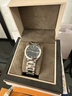 Burberry silverfärgad klocka - Stilren klocka från Burberry med silverfärgat armband i metall och svart urtavla. Klassisk design med datumvisning och tydliga indexmarkeringar. Kommer i originalask med Burberry-logga. Perfekt accessoar för dig som gillar tidlös och elegant stil.  Finns lite repor på baksidan och batterier behövs bytas !