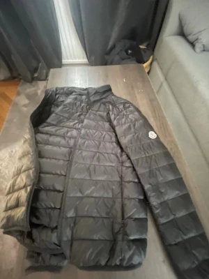 Svart dunjacka från Moncler - Snygg svart dunjacka från Moncler med klassisk quiltad design och Moncler-logga på ärmen. Jackan har hög krage och dragkedja framtill. Perfekt för kalla vinterdagar och ger en stilren look. Storlek M men passar S
