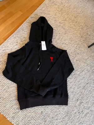 Svart hoodie från Ami Paris med röd logga - Svart hoodie från Ami Paris med klassisk huva och dragsko. På bröstet sitter Ami Paris ikoniska röda hjärta med A-broderi. Hoodien har känguruficka och ribbade muddar. Perfekt för en chill och stilren look.