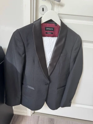 Kostymbyxor från Opposuits, grå - Snygg kostym från Opposuits med svarta satindetaljer på kavajens slag. Kostymen är helfodrad och byxorna har en beige insida med röd detalj. Använd en gång. Byxorna och kavaj är ett set och skjortan kommer seperat med också