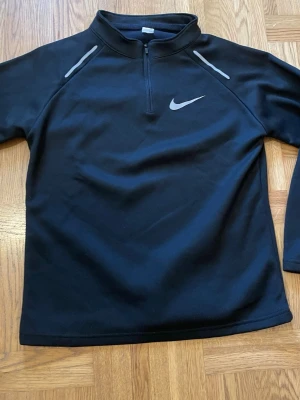 Svart Nike träningströja med dragkedja.  - Svart långärmad träningströja från Nike med halv dragkedja framtill och gråa detaljer vid axlarna. Tröjan har en hög krage och är tillverkad i ett mjukt, stretchigt material som passar perfekt för sport och träning. Den passar storlek S och kanske M.