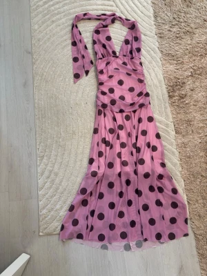 Lång prickig klänning i rosa och svart - Säljer en långklänning i rosa med stora svarta prickar. Klänningen har halterneck med knyt i nacken och en figurnära passform upptill. Materialet känns lätt och flowy, perfekt för sommarens tillfällen. Snygg och trendig look med retrovibbar.