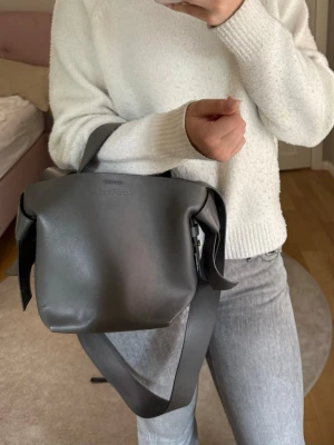 Grå axelväska från Acne Studios  - Snygg grå axelväska från Acne Studios i slätt läder med bred axelrem och cool knutdetalj på sidorna. Väskan har en minimalistisk design och är perfekt för dig som gillar stilrena accessoarer med en twist. Köpte väskan för några år sedan men ser ut som ny fortfarande då den bara har använts fåtal gånger. 