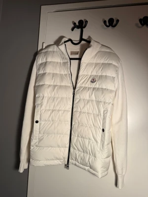 Vit cardiganjacka från Moncler - Stilren vit cardiganjacka från Moncler med quiltad framsida och stickade ärmar och rygg. Jackan har dragkedja, två fickor med knappar och Moncler-logga på bröstet. Perfekt för lager-på-lager och snygg till många outfits.