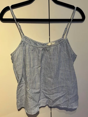 Randig linnetopp från H&M - Säljer en ljusblå och vit randig linnetopp från H&M med tunna axelband och avslappnad passform. Perfekt för varma dagar och enkel att matcha med jeans eller shorts. Materialet känns svalt och luftigt.