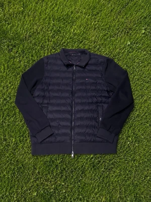 Svart pufferjacka från Tommy Hilfiger - Snygg svart pufferjacka från Tommy Hilfiger med quiltad framsida och släta ärmar. Jackan har dragkedja hela vägen, klassisk krage och diskret logga på bröstet. Perfekt för kalla dagar och enkel att matcha med olika outfits.