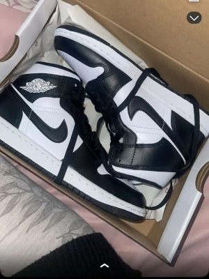 Svarta och vita Air Jordan 1 High - Säljer ett par klassiska Air Jordan 1 High sneakers i svart och vitt läder. Skorna har den ikoniska Nike Swoosh-loggan på sidorna och Air Jordan Wings-märket vid ankeln. Perfekta för dig som gillar streetwear och vill sticka ut med en tidlös modell.