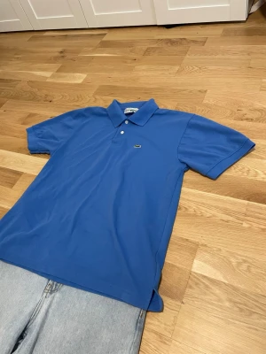 Blå pikétröja från Lacoste - Snygg blå pikétröja från Lacoste med klassisk krage och två knappar framtill. Tröjan har korta ärmar och den ikoniska krokodilloggan på bröstet. Perfekt för en avslappnad och stilren look.