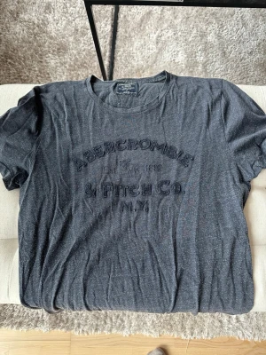 Mörkblå Abercrombie & Fitch t-shirt - Snygg mörkblå t-shirt från Abercrombie & Fitch med rund halsringning och tryckt logga framtill. Tillverkad i mjukt bomullsmaterial som känns skönt mot huden. Perfekt för en avslappnad och stilren look.