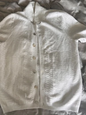 Vit kortärmad kofta från Biset - Stilren vit kofta från Biset med korta ärmar och knappar framtill. Koftan har en klassisk krage och är tillverkad i ett lätt, strukturerat material som ger en snygg textur. Perfekt att slänga över en topp för en clean look.