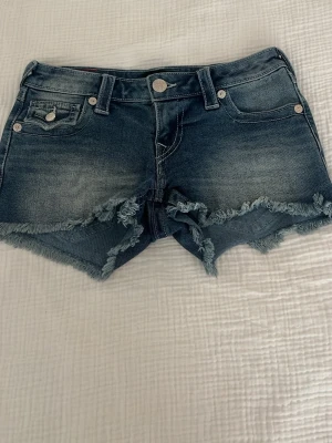 Blåa denimshorts Joey Cut-Off low waist - Snygga blåa denimshorts från True Religion, modell Joey Cut-Off. Shortsen har låg midja, fransiga kanter och klassisk femficksdesign. Perfekta för sommaren och ger en avslappnad vibe.